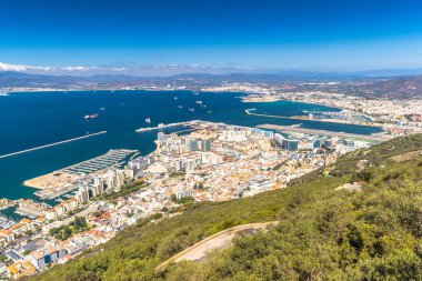 Cebelitarık, İngiliz Denizaşırı Bölgesi ve İber Yarımadası 'ndaki şehir. Sahil şehri manzarası bir tepenin üstünde, limana ve denize bakıyor, açık mavi gökyüzünün altında dağlar var..