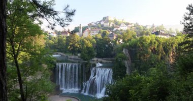 Bosna-Hersek 'in Jajce kasabasındaki Pliva Şelalesi, Avrupa. Bereketli yeşillik tarafından çerçevelenmiş manzaralı çağlayan su, arka planda temiz bir gökyüzünün altında yerleşmiş tarihi bir kasaba..