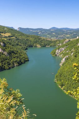 Arkasında açık mavi gökyüzü ve tepeleri olan yemyeşil bir nehir vadisi.