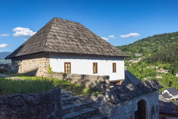 Taş temeli, beyaz duvarları ve klasik ahşap kiremitli çatısı olan büyüleyici bir Balkan evi. Ev yemyeşil dağlardan oluşan bir zemin ve açık mavi bir gökyüzüne karşı kurulmuş..