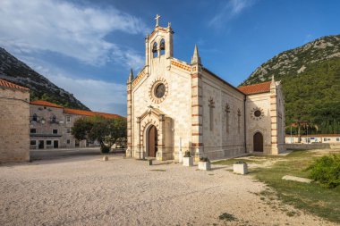 Mavi gökyüzünün altında, haçla süslenmiş eski bir kilise, çakıl tarlalarının arasında duruyor. Arkaplanda, terracotta çatısı ve gür, ormanlı dağı olan bir bina sahnenin sükunetini artırıyor..