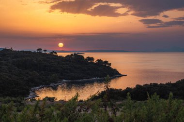 Hırvatistan, Avrupa 'nın güney Dalmaçya' daki Peljesac yarımadasındaki deniz kıyısında. Sahil boyunca gün batımının manzarası. Gökyüzünde turuncu renkler, sakin okyanus sularına ve yemyeşil sulara yansıyor..