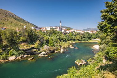 Mostar kasabası, Bosna-Hersek, Avrupa 'daki Neretva Nehri. Nehir bir Avrupa kasabasından akar. Bereketli yeşillik, parlak mavi gökyüzünün altında arka planda dağlarla çevrili binaları sarıyor..