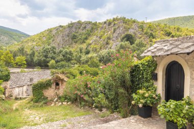 Resimli bir sahne, taştan binaların yemyeşil yamaçlara karıştığını gösteriyor. Kemerli karanlık bir kapı çiçek açan bitkilerin yanında duruyor. Taştan yollar ve çatılar kırsal çekiciliğe ekleniyor..
