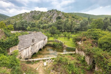Bosna-Hersek, Avrupa 'daki Blagaj köyündeki eski taş ev. Çiçekli yeşillik ve manzaralı manzarayla çevrili küçük bir nehrin üzerinde kemerli açıkları olan tarihi taş bina..