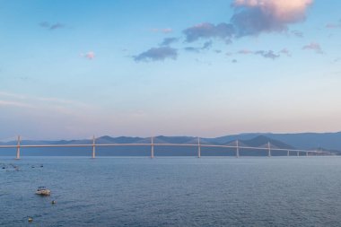 Hırvatistan, Avrupa 'daki Peljesac Yarımadası' na giden Peljesac Köprüsü. Geniş süspansiyon köprüsü sakin su boyunca uzanır, arka planda küçük bir tekne ve dağ silueti bulunur..