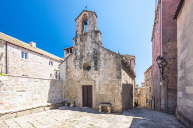 Bu resim, tarihi bir Avrupa kasabasındaki yıpranmış taş bir şapeli gösteriyor. Güneşli sahne mimari ayrıntıları, bir çan kulesi ve büyüleyici bir kaldırım taşı karesini gözler önüne seriyor. Bölge davetkar görünüyor..