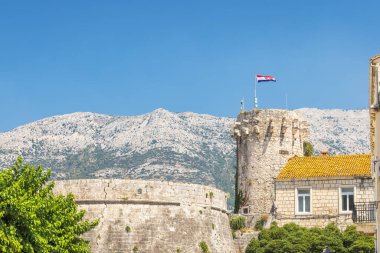 Hırvatistan, Avrupa 'daki Adriyatik Denizi' nin Korcula adasındaki Korcula kasabası. Üzerinde mavi gökyüzünün altında dağlık bir manzarayla desteklenmiş bir bayrak bulunan taş kale kulesinin yer aldığı pitoresk kıyı kasabası..