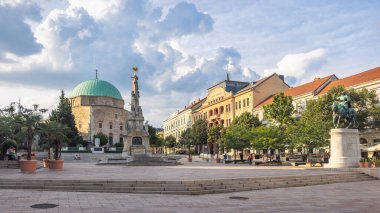 Pecs, Macaristan - 13 Temmuz 2024: Szechenyi Meydanı. Bulutlu bir gökyüzü altında yeşil kubbeli bir cami, heykeller ve tarihi binalar barındıran şehir manzarası kültürel simgeleri destekliyor..