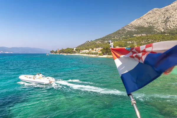 Orebiç, Hırvatistan - 11 Temmuz 2024: Peljesac Yarımadası 'nın deniz kıyısı. Turkuaz su, bereketli tepeler, bir motorlu tekne ve açık mavi gökyüzünün üzerinde dalgalanan Hırvat bayrağının dalgalandığı pitoresk bir kıyı sahnesi.