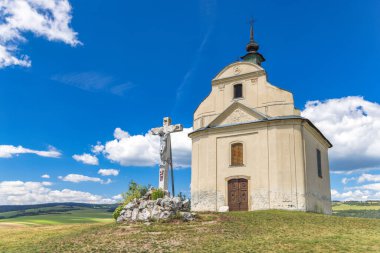 Chapel on a travertine hill of The Siva Brada in Spis region in Slovakia, Europe. Chapel and cross stand on a hill in sunlight, under a blue sky with fluffy clouds, overlooking a green landscape.