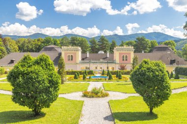 Hodkovce Mansion in Zehra village in Slovakia, Europe. Stately manor house nestled in a lush green park with a mountain backdrop under a clear blue sky filled with fluffy white clouds.