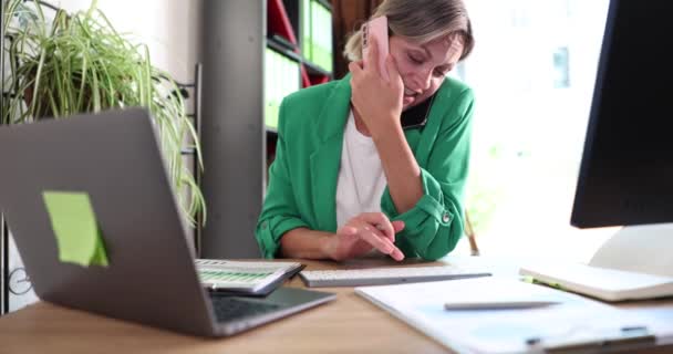 严肃的年轻女工正在电话交谈 并在办公室使用带有应用程序的笔记本电脑 Multitasking Manager一边打电话给客户一边在笔记本电脑上工作 — 图库视频影像