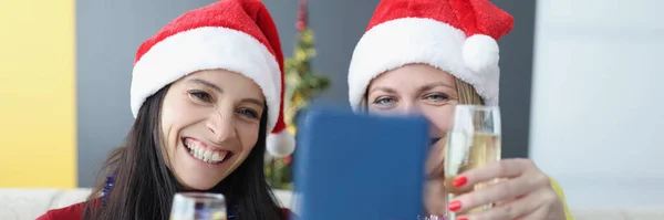 Two women in santa hats drinking champagne and taking selfie on cell phone at home. Removed merry christmas and happy new year greetings concept