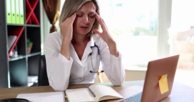 Ofiste dizüstü bilgisayarın önünde baş ağrısı ve migrenle oturan kadın doktor. Tıbbi personelin duygusal ve psikolojik stres kavramı