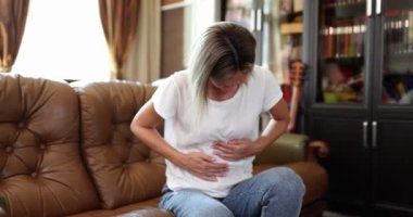 Karnına dokunan kadın karın ağrısından muzdariptir ve kendini kötü hisseder. Evdeki koltukta oturan karın ağrısı çeken bir kadın.