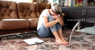Kadın, bedenin maasasını ölçmek için terazinin yanında oturmuş ağlıyor. Aşırı kilolu depresyon ve stres