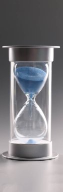 Close-up of hourglass with blue sand on gray background. Sand flowing through the bulb of sandglass. Invertible device, retro symbol of time