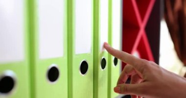 Kadın elleri raftaki klasörlere dokunmuş, yakından, yavaş çekimde. Mali belge yönetimi, arşiv