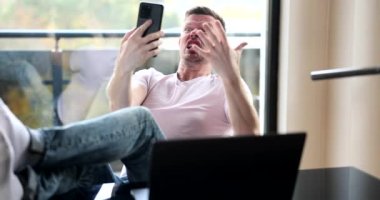 Kızgın adam masada oturuyor ve cep telefonu 4K yavaş çekim ekranına bağırıyor. Uzak çalışma kavramının zorlukları 
