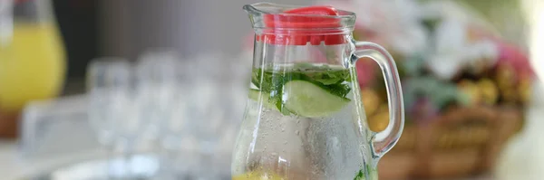 Glass jug of detox water with lemon and cucumber standing on table closeup. Healthy food concept