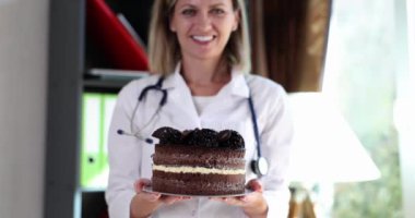 Doktor diyetisyen mutlu gülümsüyor ve elinde çikolatalı pasta tutuyor. Şeker ve tatlı kalori pastaları vücuda zarar verir.