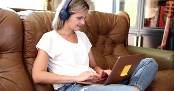 ヘッドフォンの女性はラップトップでソファに座っています リモートワークトレーニングと語学コース — ストック動画