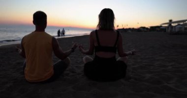 Gün batımında, bir adam ve bir kadın kumların üzerinde oturmuş meditasyon yapıyorlar, yakın plan. Sahilde yoga konsepti
