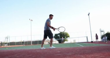 Bir erkek ve bir kadın açık tenis kortu oynuyorlar, yavaş çekimde. Popüler bir spor oyunu öğretmeyi saymazsak, oyuncular