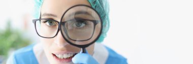 Portrait of doctor in glasses looks through magnifying loupe, intern provide investigation in clinic. Medical worker use magnifying glass. Medicine concept