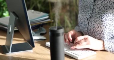 İş kadını, taşınabilir hava nemlendiricisinin yanında bilgisayar klavyesinde çalışıyor. 4K film yavaş çekim. Oda konseptinde rahat bir mikro iklim yaratmak için ev aletleri