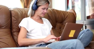 Kulaklıklı gülümseyen kadın kanepeye oturur ve dizüstü bilgisayarda webinar izler. Kulaklıklı mutlu genç kız oturma odasındaki koltukta bilgisayar kullanıyor.
