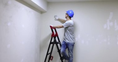 Miğferli erkek müteahhit duvara alçı yapıştırma kalitesini ölçüyor ve ağır çekimde jest gösteriyor. Dairelerin yüksek kaliteli onarımı ve dekorasyonu