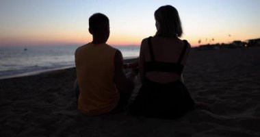 Couple in love watching sunset on beach together. Silhouette of people in love from behind sitting enjoying view of sunset sea
