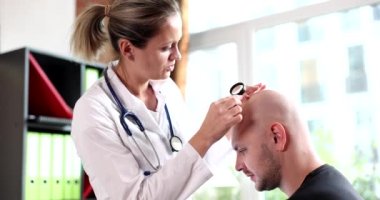 Female trichologist with magnifying glass checks bald head of adult man. Trichologists consultation