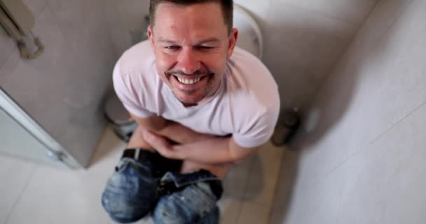 幸せな笑顔の男は自宅のバスルームでトイレに座っている 鼓腸は診断と治療を引き起こす — ストック動画