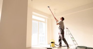 Builder manually paints the ceiling inside house and building. Repairman using a white roller with long handle at construction site