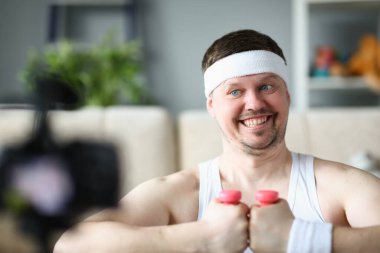 Smiling man is holding dumbbell and looking into camera. Athlete blogger and remote home fitness concept