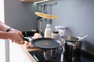 Hands hold black iron pan and toss pancake into air. Cooking pancakes at home in kitchen