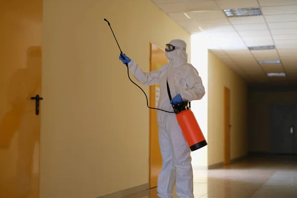 Contractor disinfects office and corridor from coronavirus COVID-19. Disinfectant in protective suit and mask sprays disinfectants in clinic