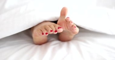 Woman legs stick out from under blanket in bed with red pedicure. Sleep problems and insomnia concept