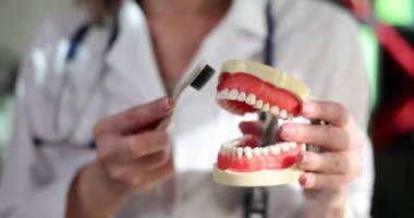 Diş hekimi, diş hekiminde dişlerini fırçalaması gerektiğini gösteriyor. Doktor, beyaz diş fırçasıyla diş temizleme hareketleri gösteriyor.