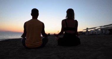 Genç bir çift gün doğumunda sahilde meditasyon yapıyor. Lotus pozisyonunda otururken