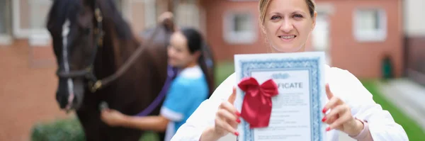 Woman veterinarian holds medical certificate on background of horse. Thoroughbred sport horses and passport concept