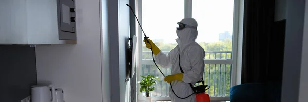 Worker in protective suit cleans room from cockroaches and rats with spray gun. Sanitary service disinfects the apartment with chemical agent