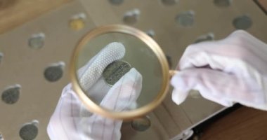 Numismatist examines coin from collection through magnifying glass. Valuation of ancient coins