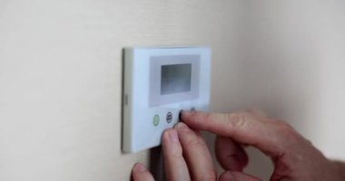 Human hands adjust room temperature on digital thermostat in apartment. Climate control and air temperature in hotel room