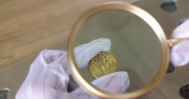 Hands in gloves hold an old coin and a magnifying glass, close-up. Estimated value from an antique dealer