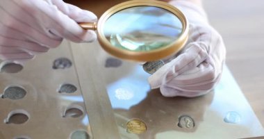Numismatist in gloves with magnifying glass is examining collection of coins. Passionate collector of old coins