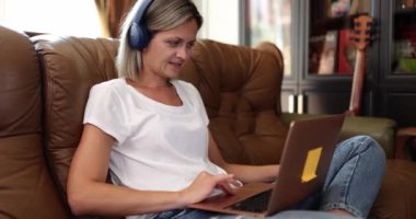 Female freelancer sits on sofa and uses laptop and headphones. Video chat with friends watching movies or teleworking teleworking